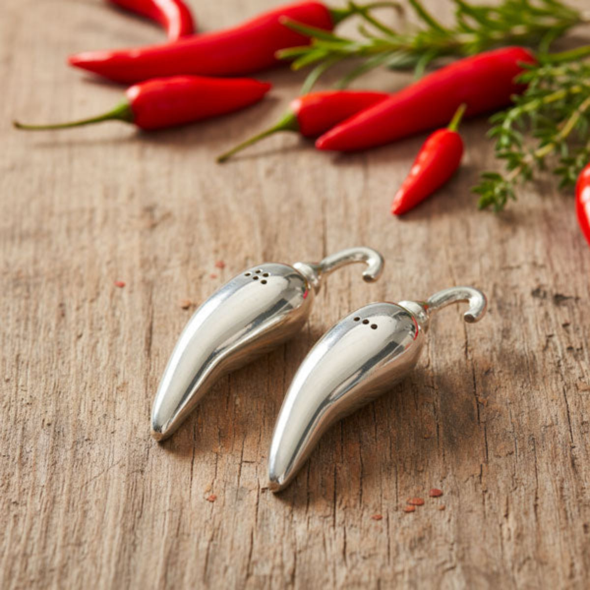 Two metallic chili pepper-shaped salt and pepper shakers on a rustic wooden surface, with whole red chili peppers and green herbs blurred in the background.