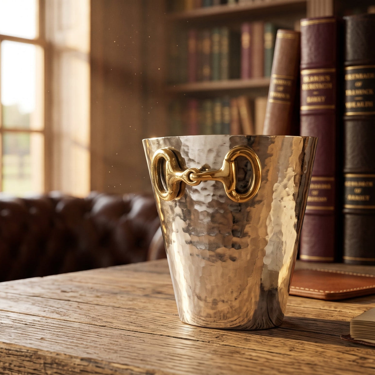 A hammered metallic bucket with horsebit handles sits on a rustic wooden table in a warm, sunlit library or study room with bookshelves and a leather chair in the background.