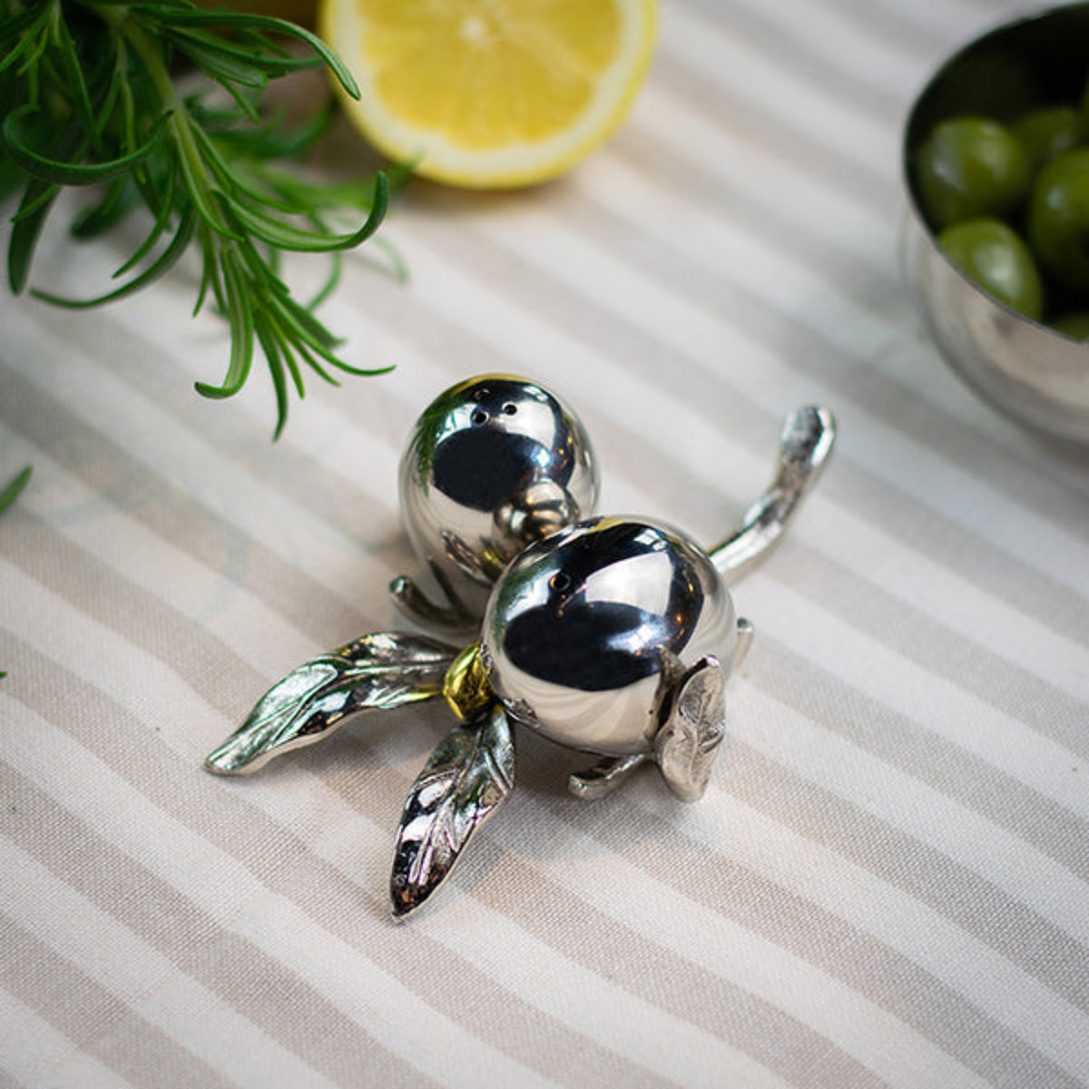 A polished silver decorative object shaped like two small fruits with leaves, placed on a striped fabric surface with a lemon slice, greenery, and a bowl of olives in the background.