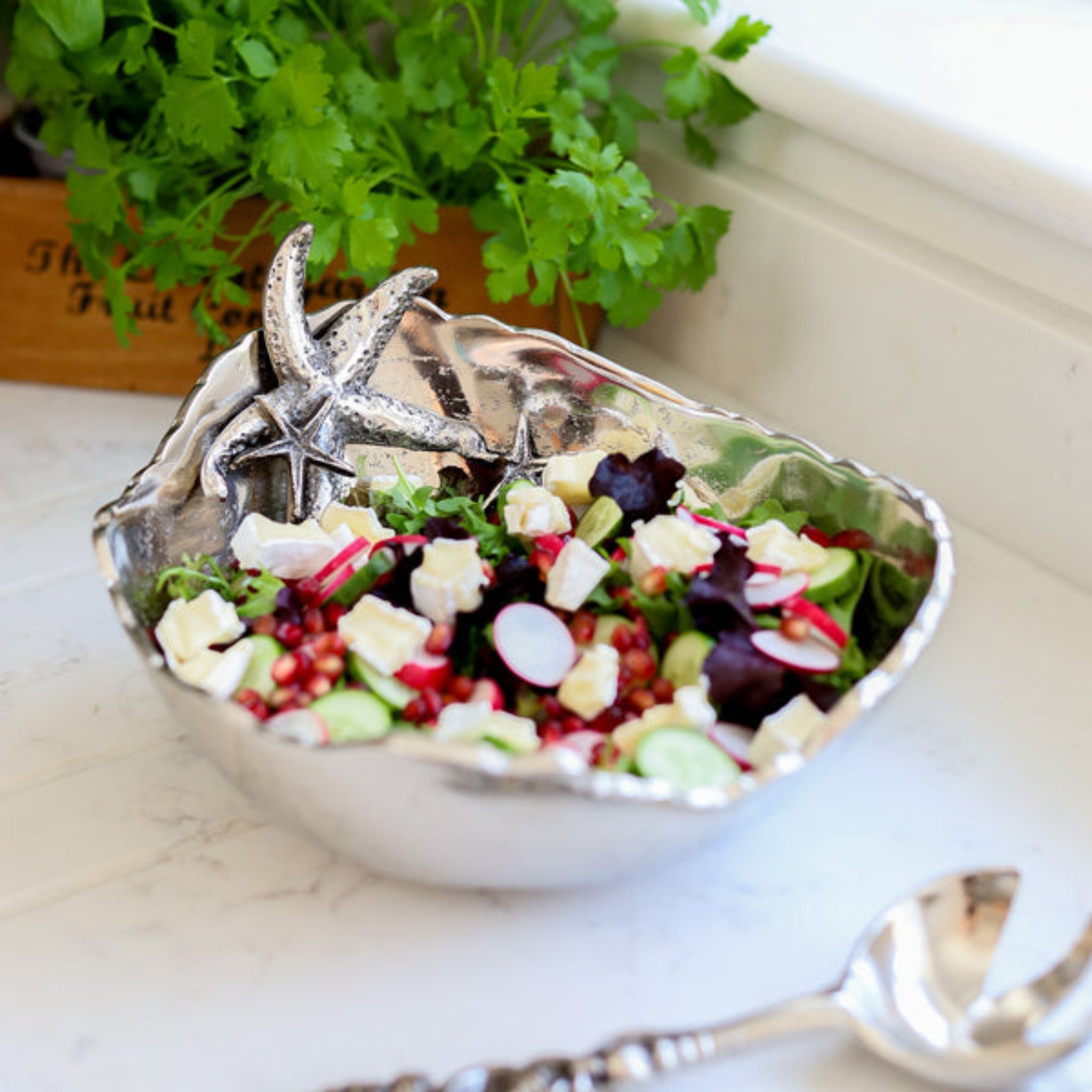 Silver bowl with a starfish design containing a salad on a marble surface with a plant in the background.
