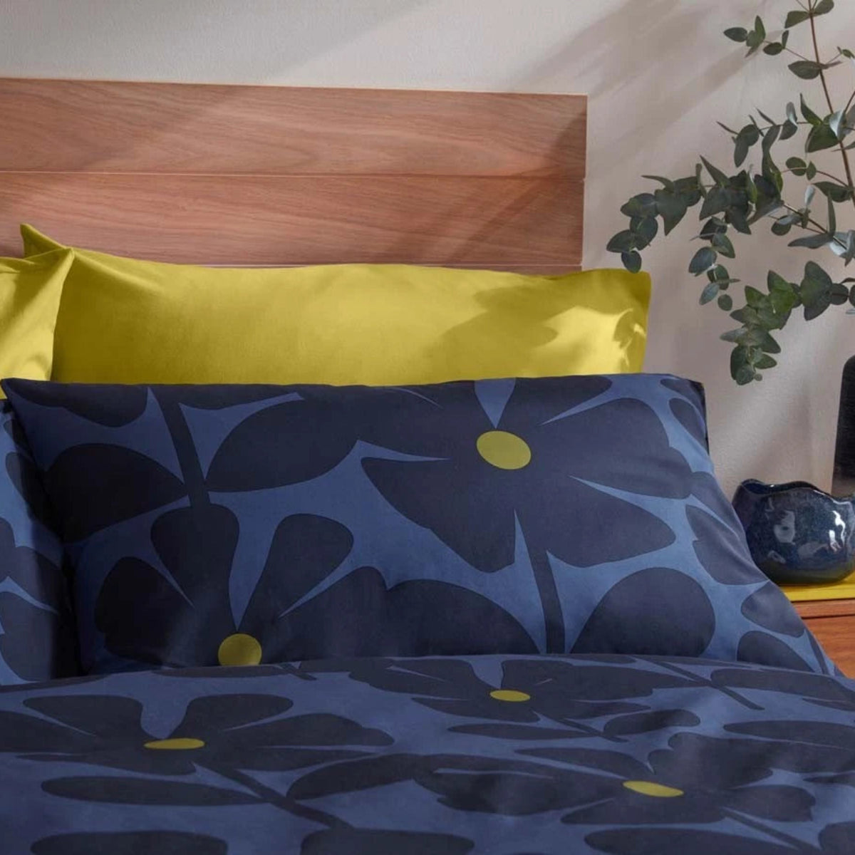 A styled bed featuring a navy blue and blue floral pattern duvet cover and matching pillowcases with yellow flower centers, layered with solid yellow pillows, set against a wooden headboard and a plant.