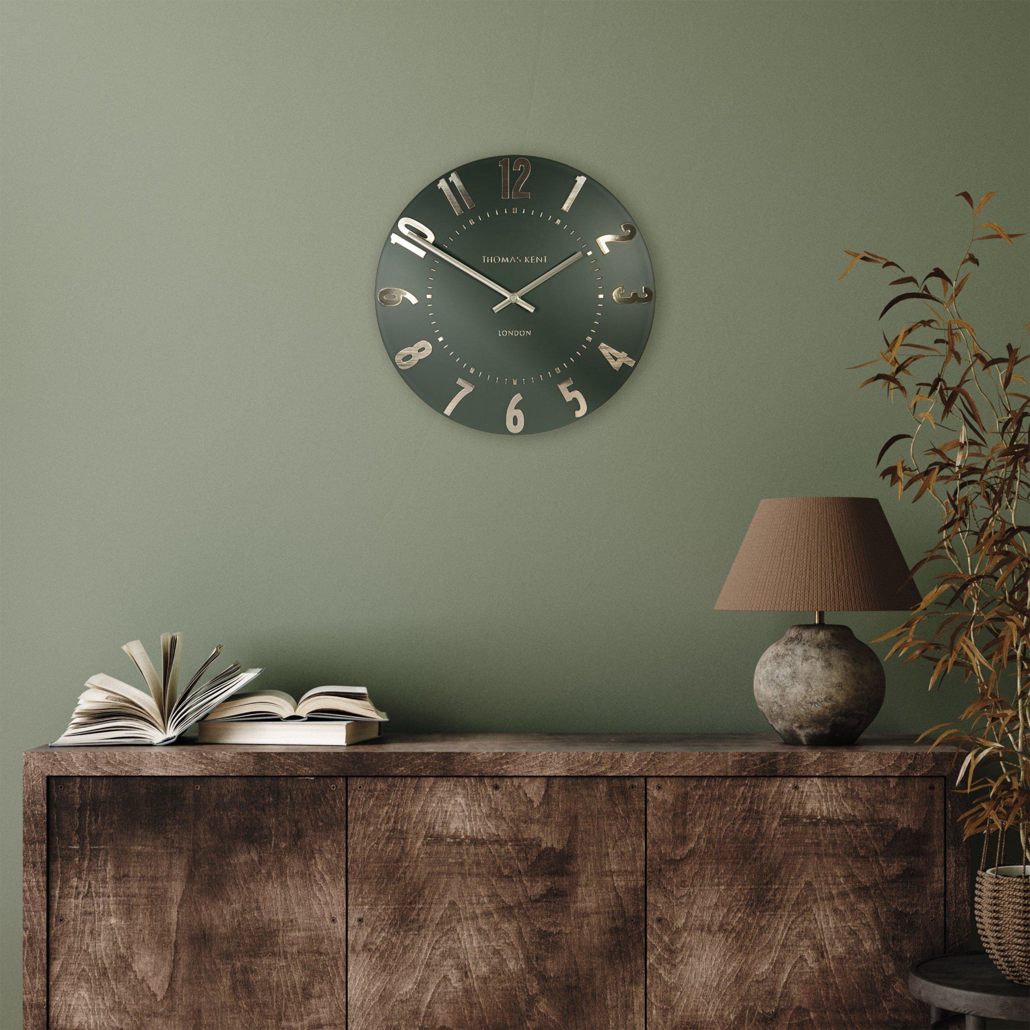 Wall clock on a green wall above a wooden cabinet with books and a lamp.