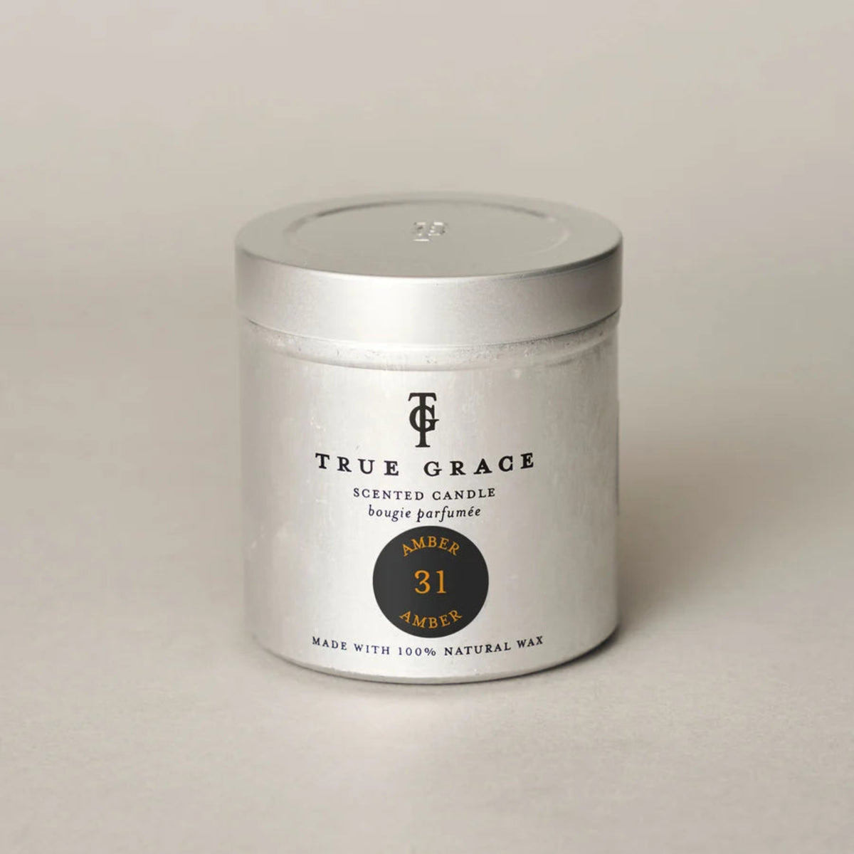 A silver metal tin candle with a matching lid, featuring a white label with black text and a circular amber-colored scent badge. The brand &