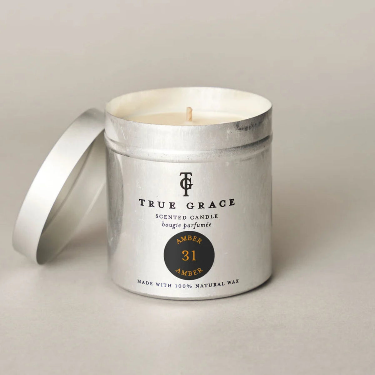 A silver metal scented candle tin with its lid leaning against it on a light gray surface. The label reads TRUE GRACE and shows amber 31 amber.