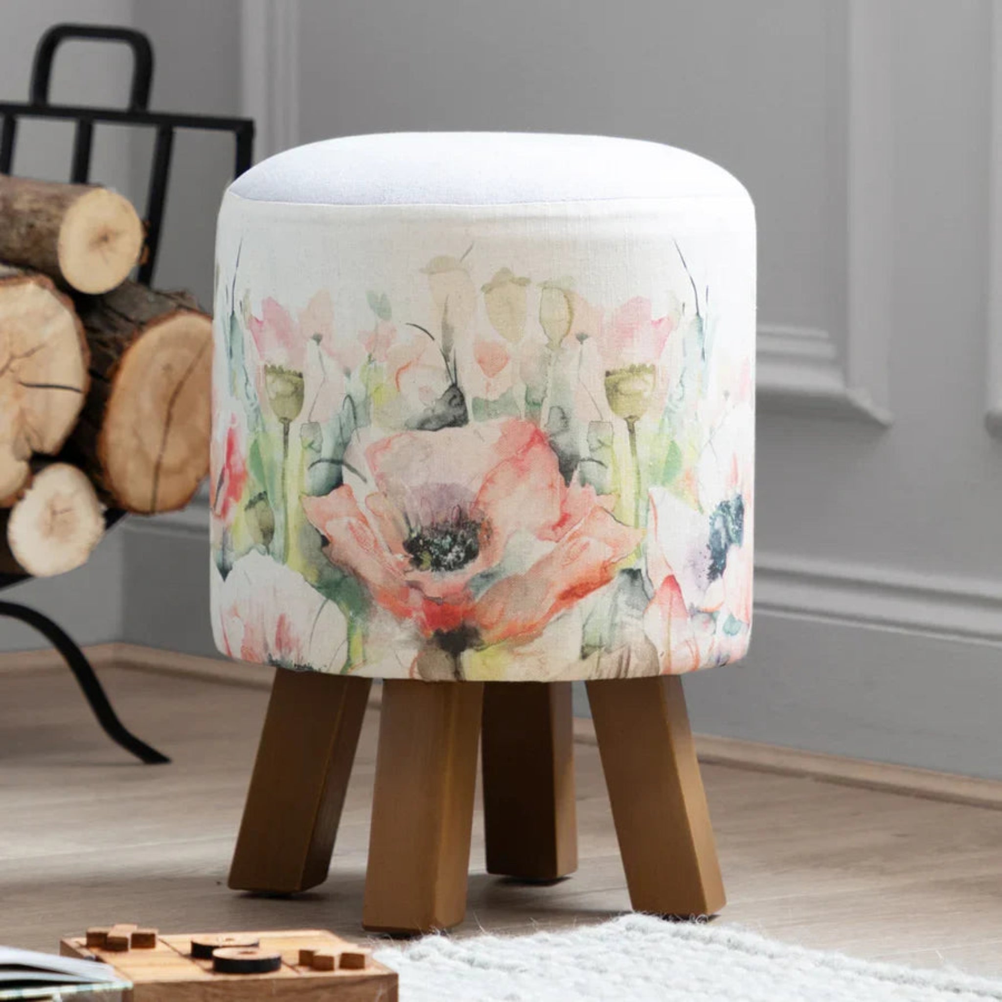 A round upholstered stool with a white top and four wooden legs, covered in fabric with a watercolor-style floral print featuring pink, peach, and green flowers, shown in a room setting.