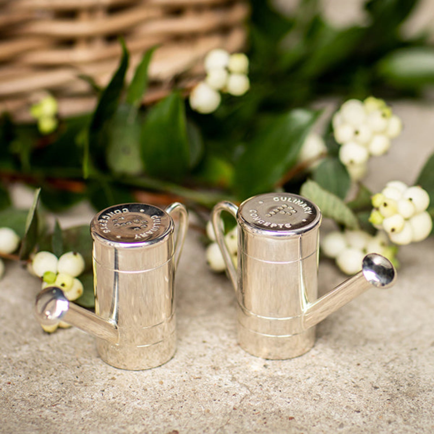 Two silver watering cans on a textured surface with greenery and flowers in the background