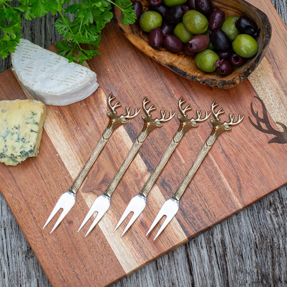 Set of four gold deer-head patterned forks on a wooden board with cheese and grapes.