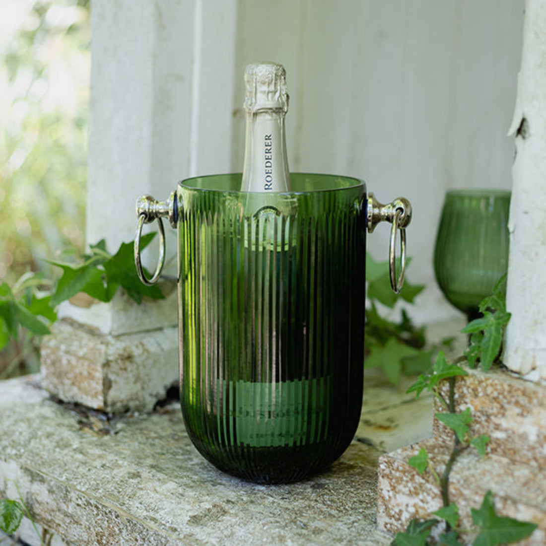Green glass ice bucket with a bottle of champagne on a stone ledge outdoors.