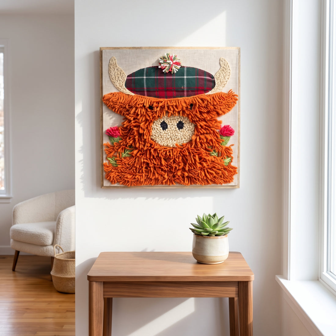 A rustic, handcrafted wall hanging featuring a fluffy orange shaggy animal face with a tartan plaid hat, displayed on a white wall above a wooden console table in a bright, modern room.