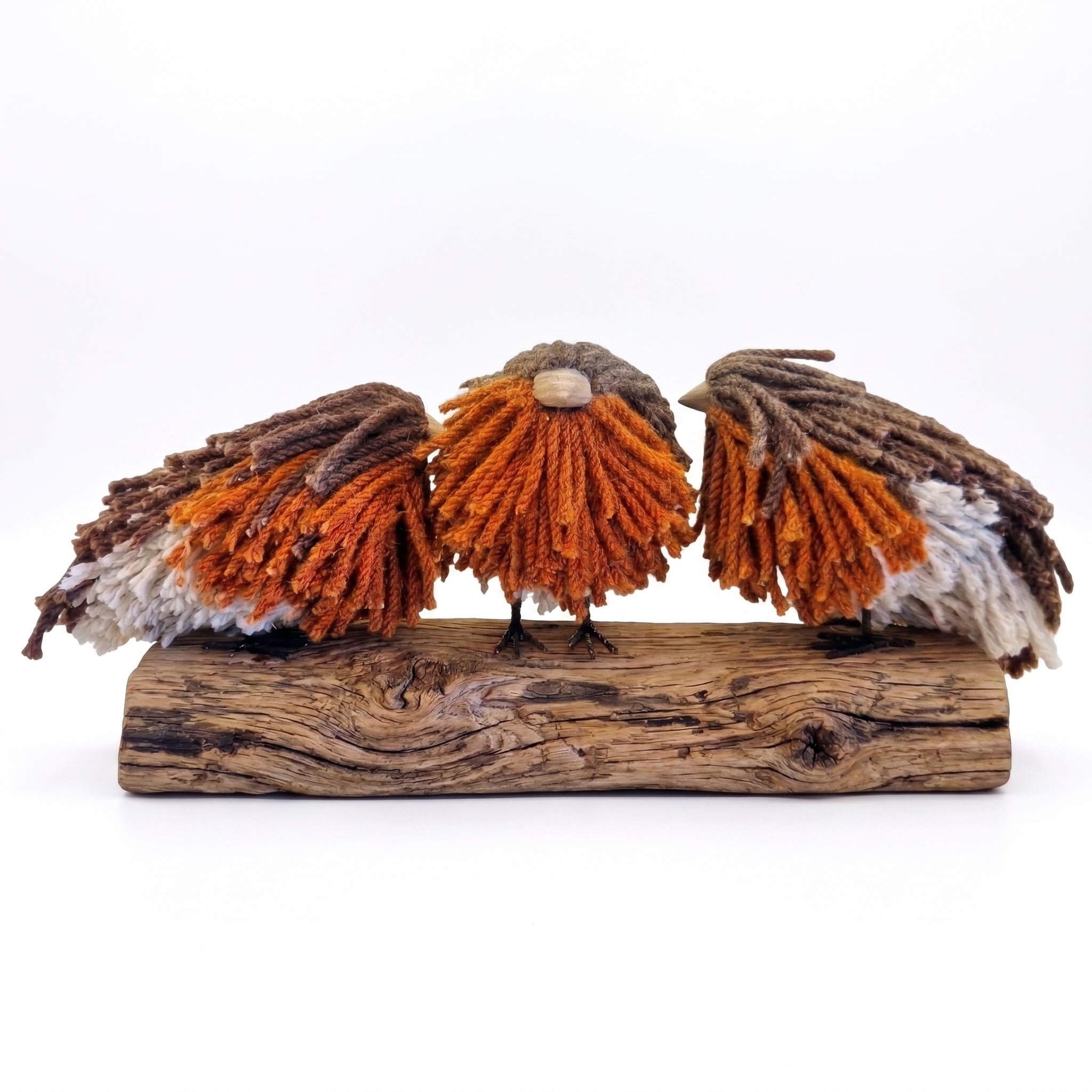 Three decorative birds crafted from orange, brown, and white yarn, perched on a rustic piece of driftwood against a plain white background.