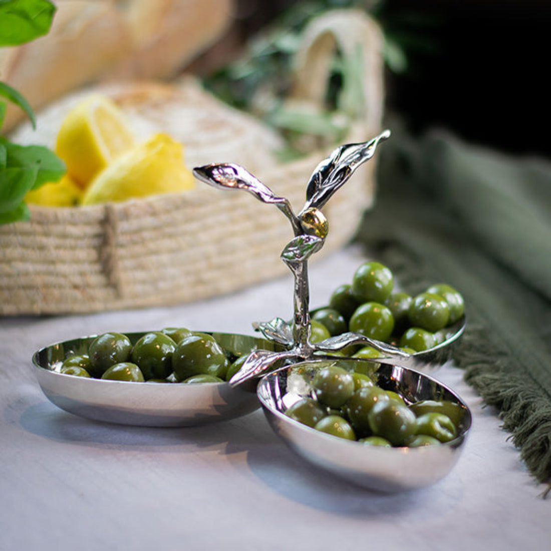 Three silver olive dishes with a decorative handle on a table with green olives.