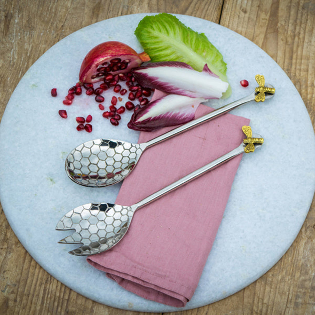 Two silver spoons with gold bee handles on a pink napkin with salad and pomegranate seeds.