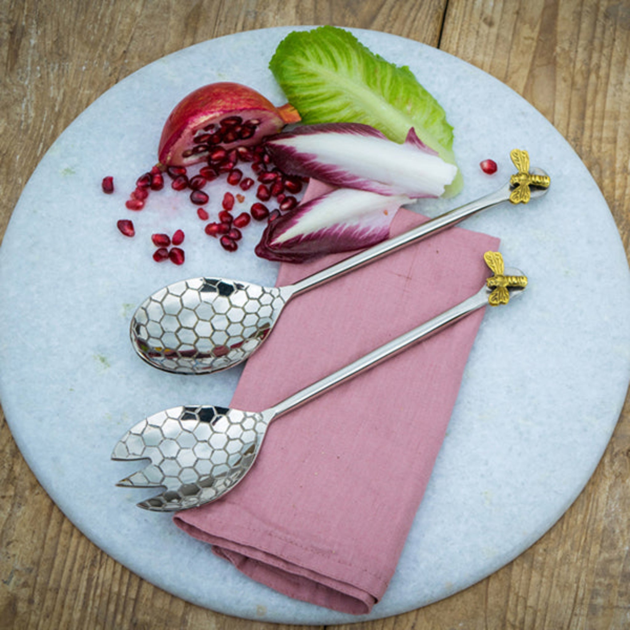 Two silver spoons with gold bee handles on a pink napkin with salad and pomegranate seeds.