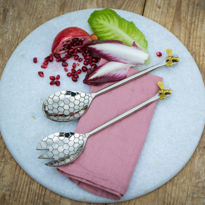 Two silver spoons with gold bee handles on a pink napkin with salad and pomegranate seeds.
