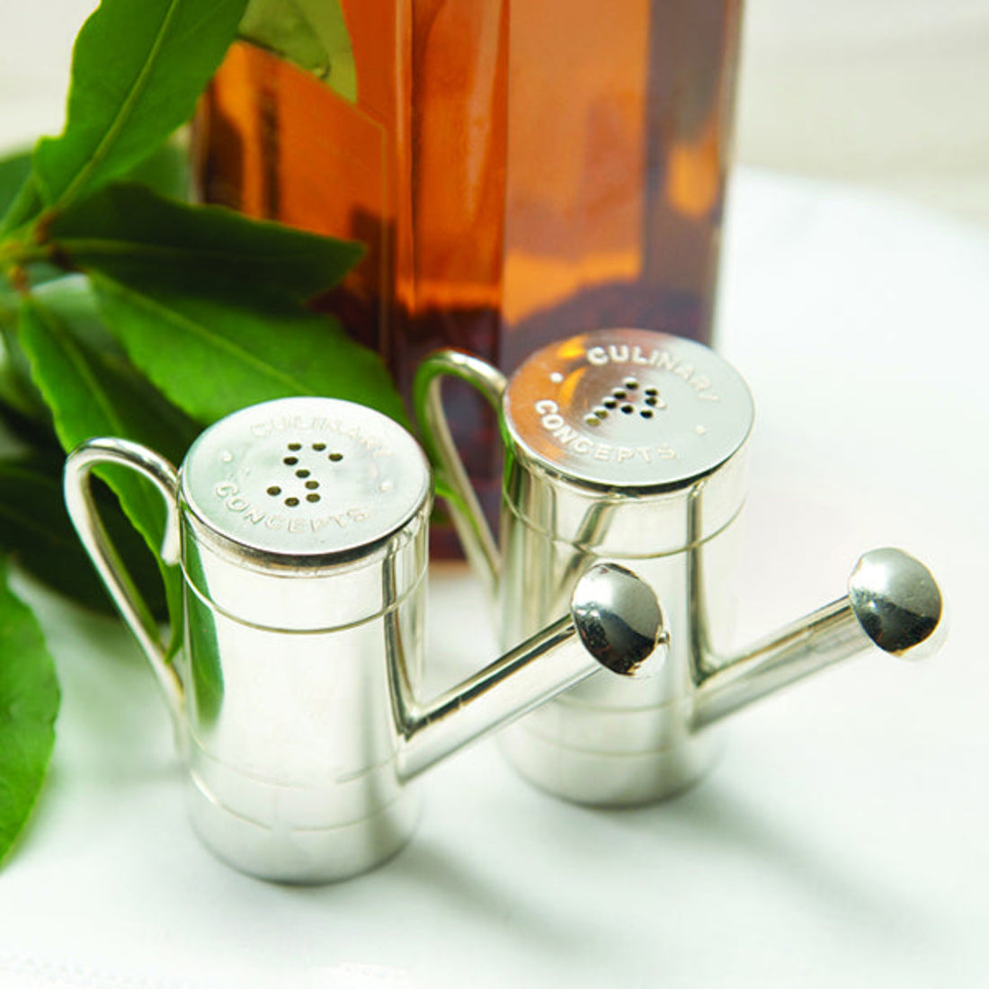 Silver salt and pepper shakers with Culinary Concepts branding on a white surface with green leaves and a bottle in the background.