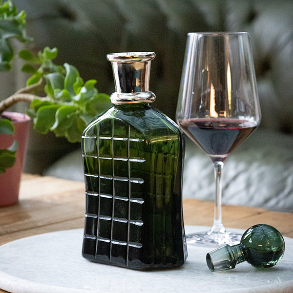 Green glass bottle with grid pattern next to a wine glass on a table with a plant in the background