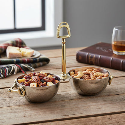 A two-bowl hammered metal snack stand with a gold-tone handle, shown on a rustic wooden table with nuts and dried fruit in the bowls, alongside a book, a glass of amber liquid, and a folded plaid blanket.