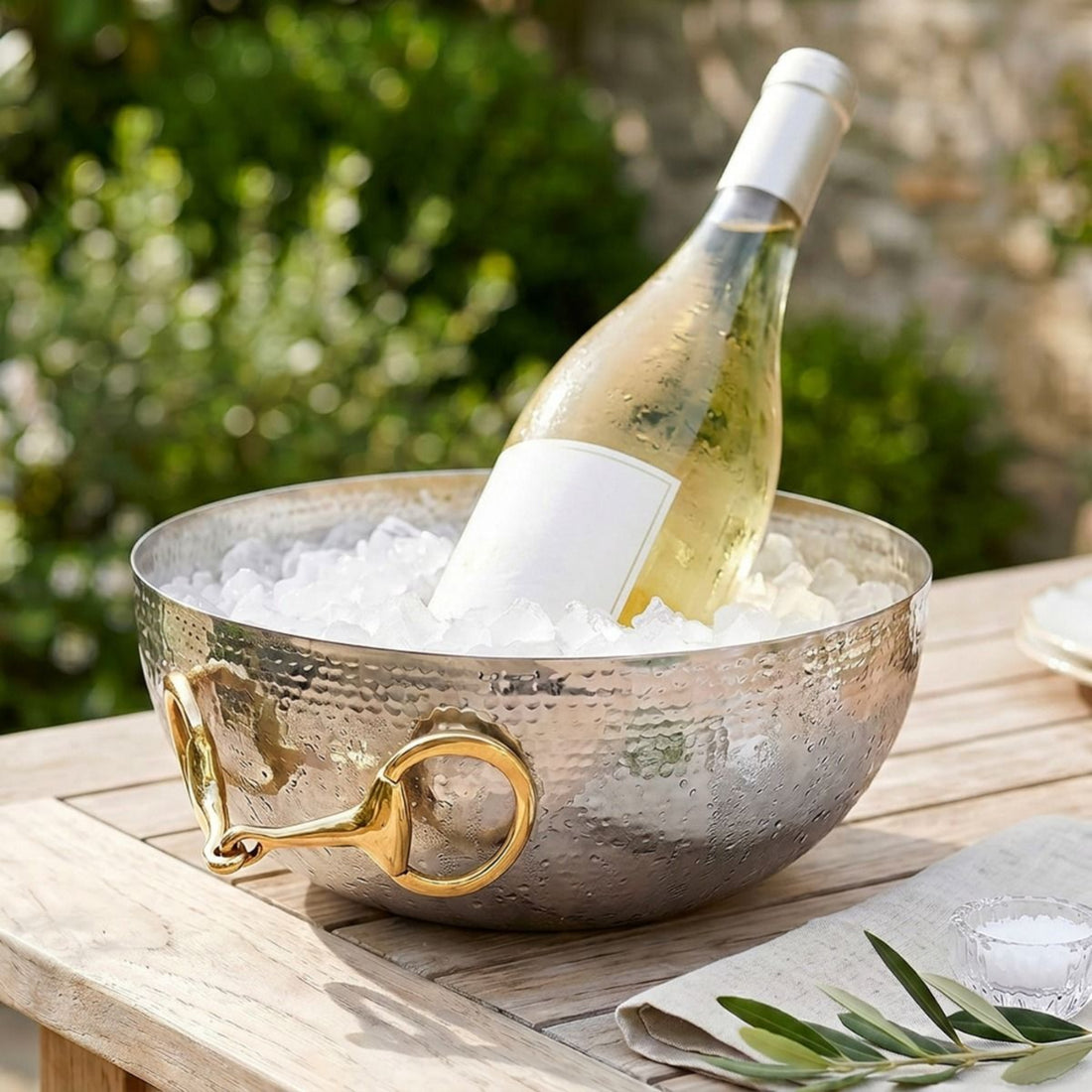 A hammered silver metal wine bucket with gold horsebit handles, filled with ice and holding a white wine bottle, set on a wooden table outdoors with green foliage in the background.