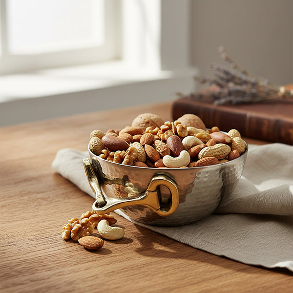 A hammered silver bowl filled with assorted nuts and almonds, set on a wooden table with a beige cloth and a blurred book in the background.