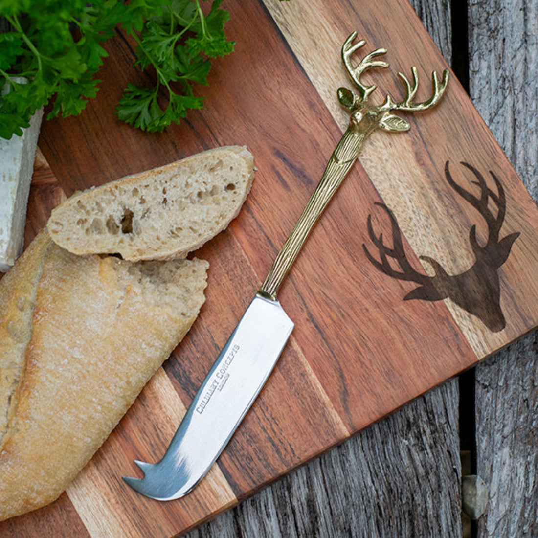 Cheese knife with deer head design on a wooden cutting board with bread and herbs.