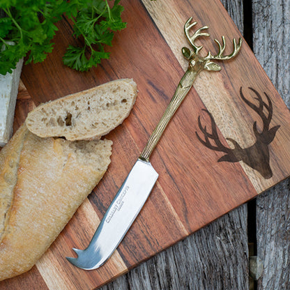 Cheese knife with deer head design on a wooden cutting board with bread and herbs.