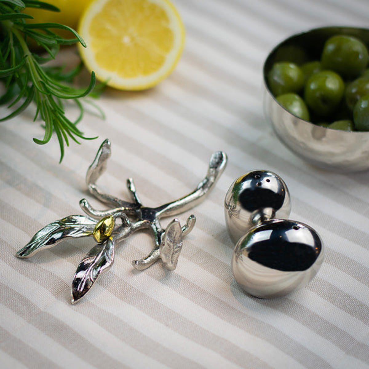 Silver-toned olive branch and olive-shaped cabinet knobs on a striped fabric surface, styled with a lemon slice, rosemary, and a bowl of green olives.