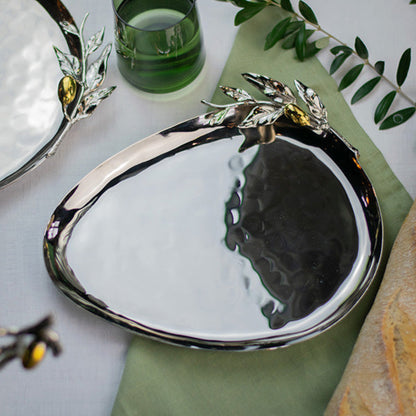 A polished silver serving platter with decorative leaf and olive branch handles, styled on a green napkin with bread and a green glass in the background.