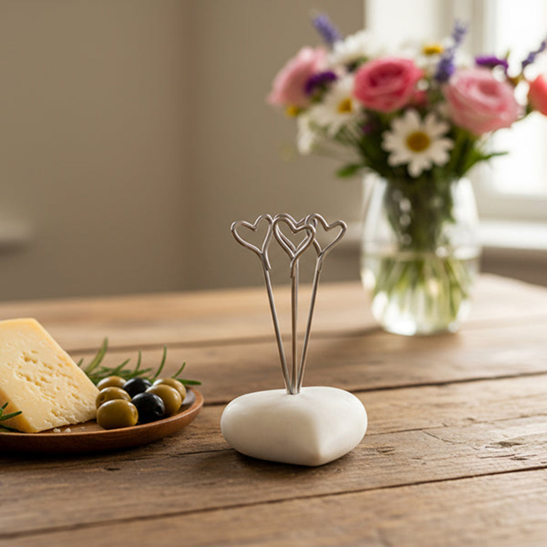 Decorative metal holder on a white stone base with a plate of cheese and olives, and a vase of flowers in the background.