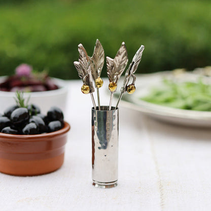 A set of decorative silver-toned metal cocktail picks shaped like leaves, each topped with a small gold bead, displayed in a small cylindrical metal holder on a white tablecloth with blurred food and greenery in the background.
