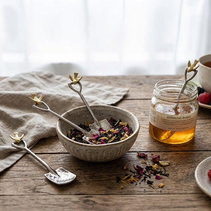 Three silver spoons with gold-toned flower handles are arranged on a rustic wooden table. One spoon rests in a ceramic bowl filled with dried flower petals and tea, while the others are placed beside it.