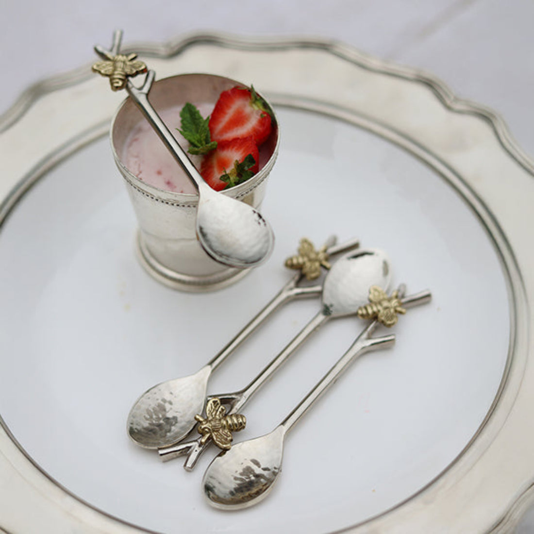 Three decorative silver spoons with gold bee handles are arranged on a white plate, alongside a small silver cup containing a pink dessert topped with a strawberry and mint.