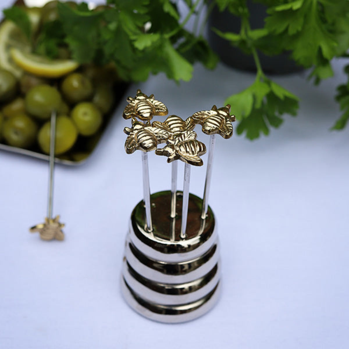 A set of gold-tone metal cocktail picks with bee-shaped tops, displayed in a striped silver and black holder on a light surface, with olives, lemon, and green herbs in the background.
