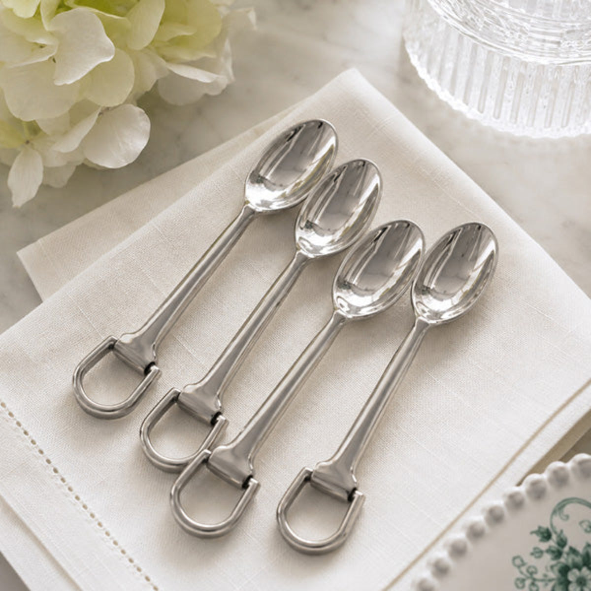Four polished silver spoons with integrated ring handles, arranged diagonally on a folded white napkin with soft floral props in the background.