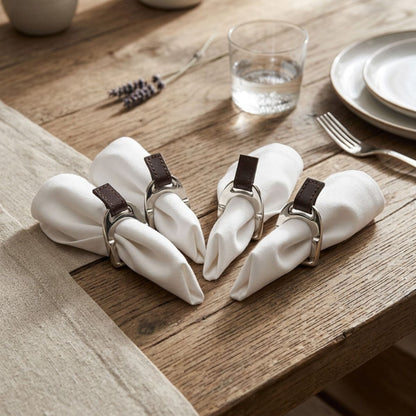 Four white cloth napkins rolled and secured with silver-toned metal rings that have dark brown leather tabs, arranged on a rustic wooden table with a glass, plates, and cutlery nearby.
