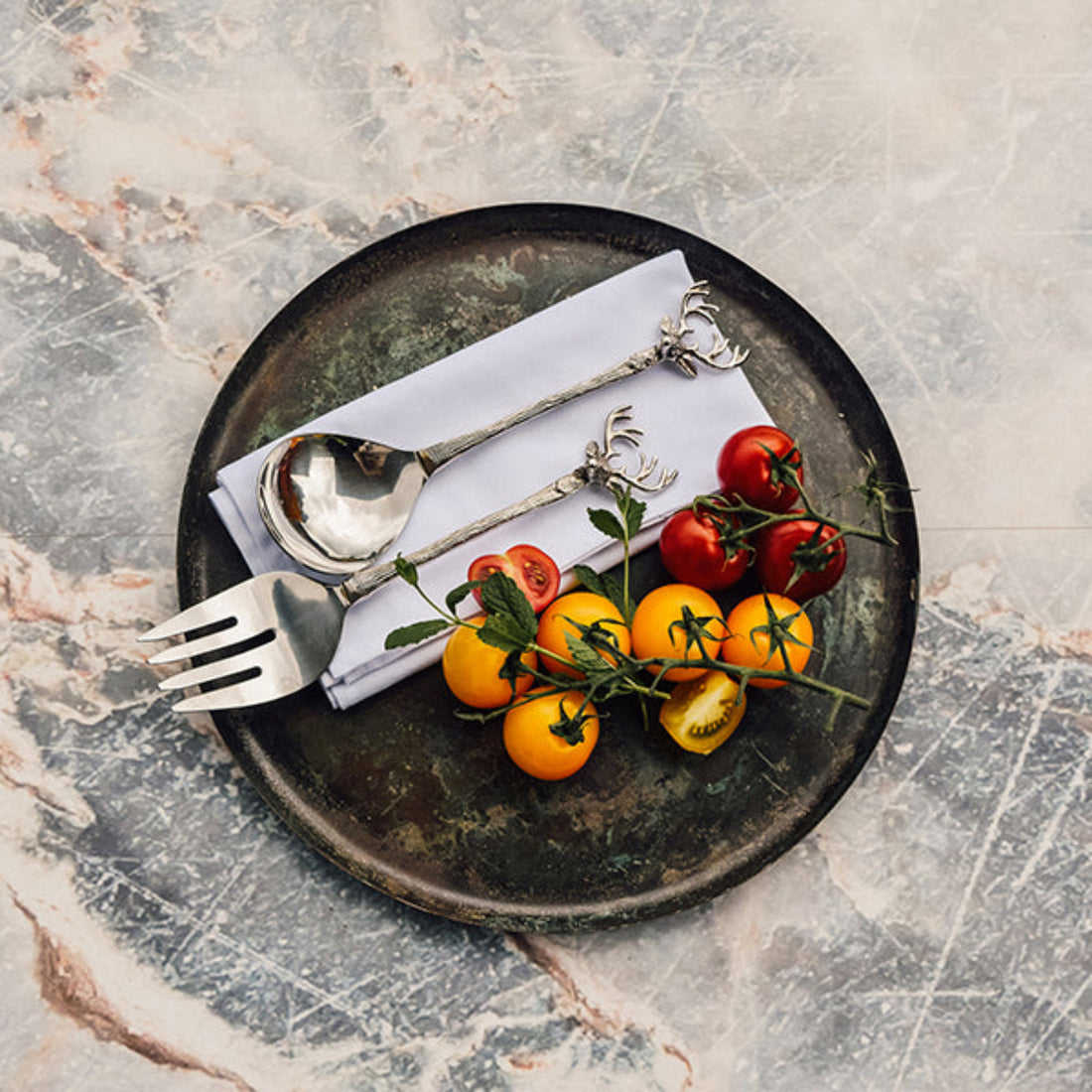 Black plate with silver fork and spoon with a stag design, white napkin, and cherry tomatoes on a marble surface