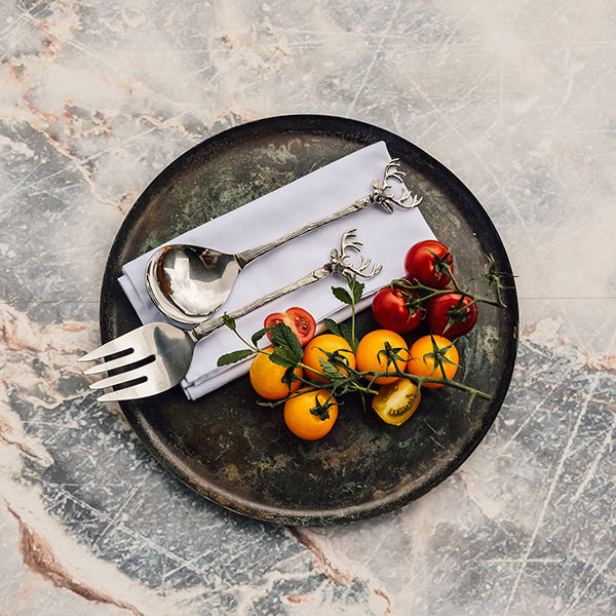 Black plate with silver fork and spoon with a stag design, white napkin, and cherry tomatoes on a marble surface