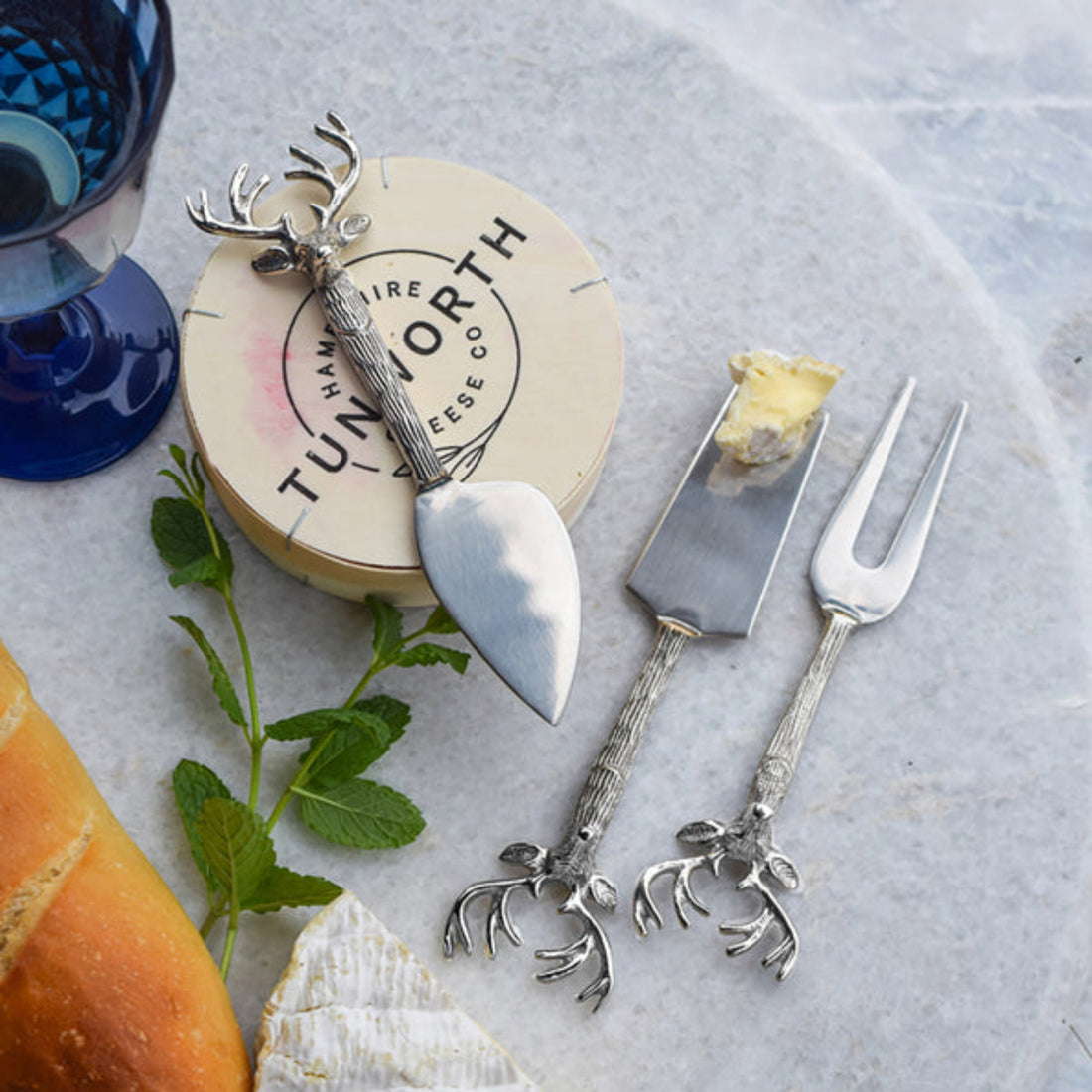 Silver cheese tools with deer antler design on a marble surface with a cheese board and bread.