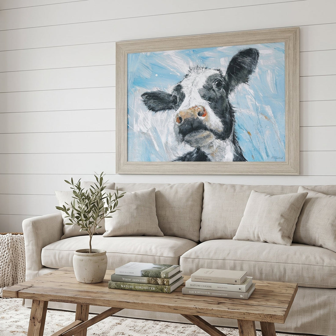 A framed canvas art print of a black and white cow painting on a light blue background, displayed on a white shiplap wall above a beige fabric sofa in a bright, modern living room.