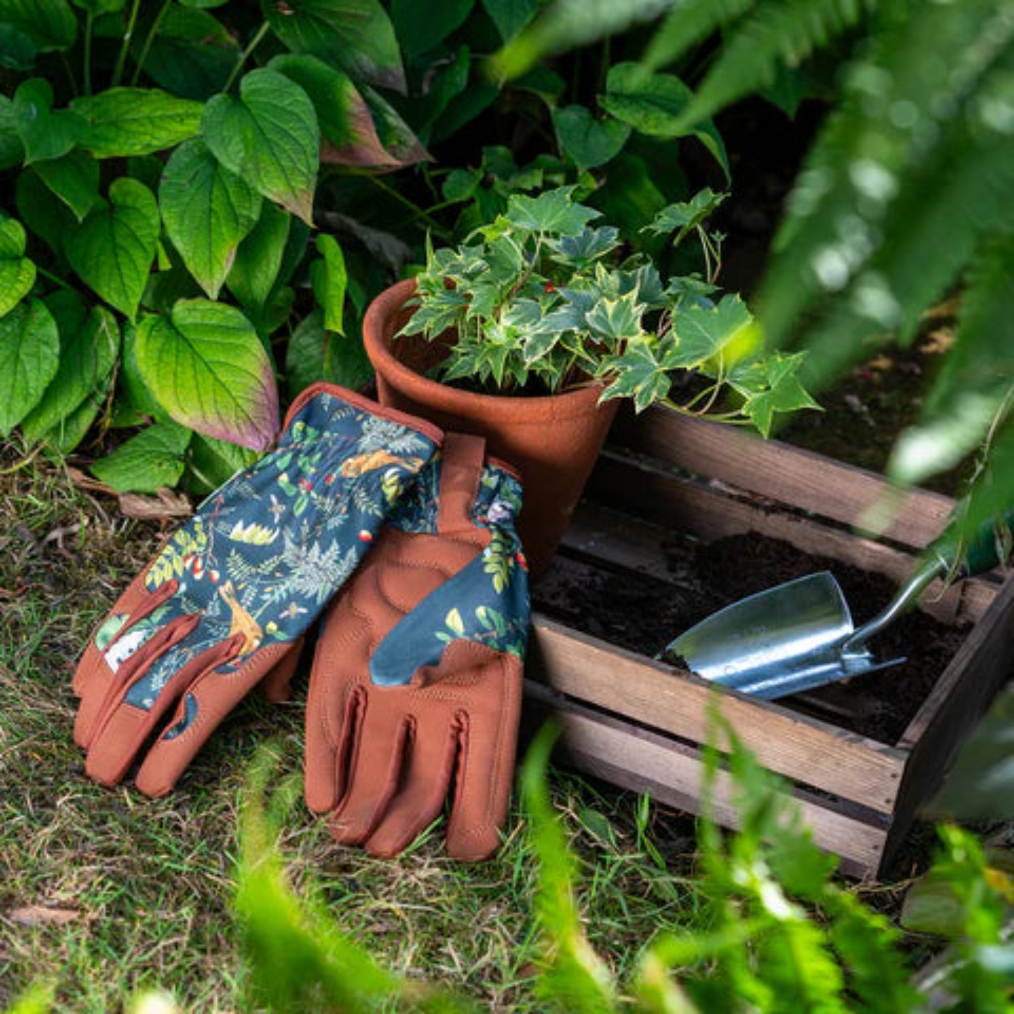 Gardening gloves and tools on a garden bed with plants and a wooden crate.
