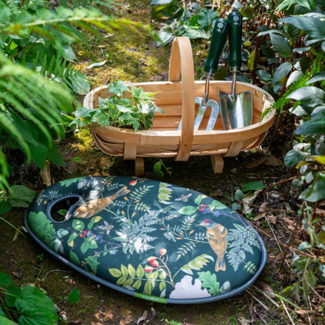 Gardening tools in a wooden basket with a decorative garden-themed cushion on the ground.