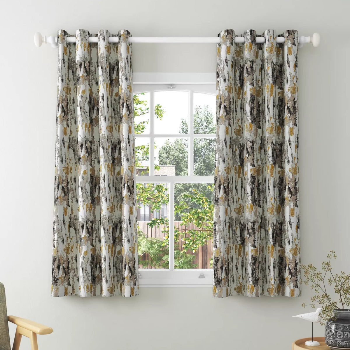 A pair of grommet-top curtains hanging on a white rod in a bright room. The curtains have an abstract pattern in gray, beige, white, and muted gold tones.