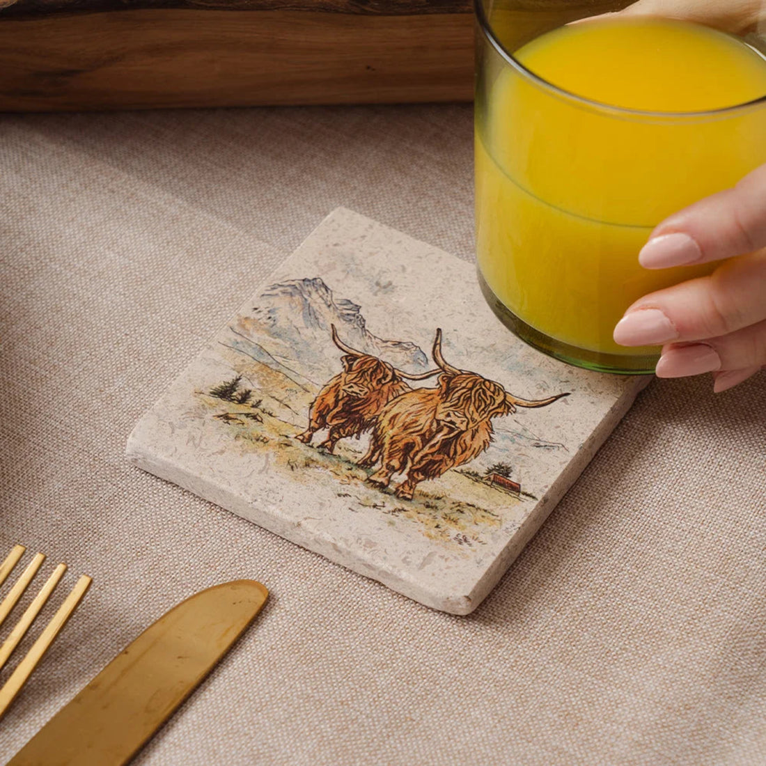 Coaster with highland cow design holding a glass of orange juice on a table.