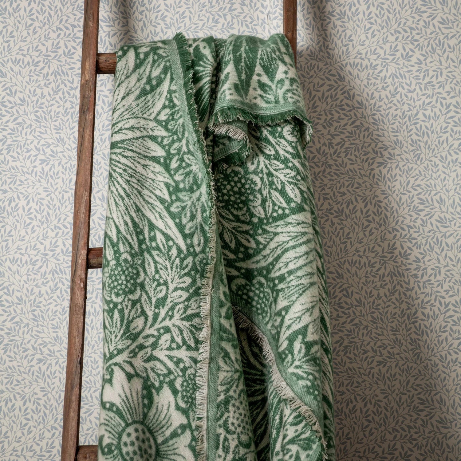 Green patterned blanket draped over a wooden ladder against a textured wall.