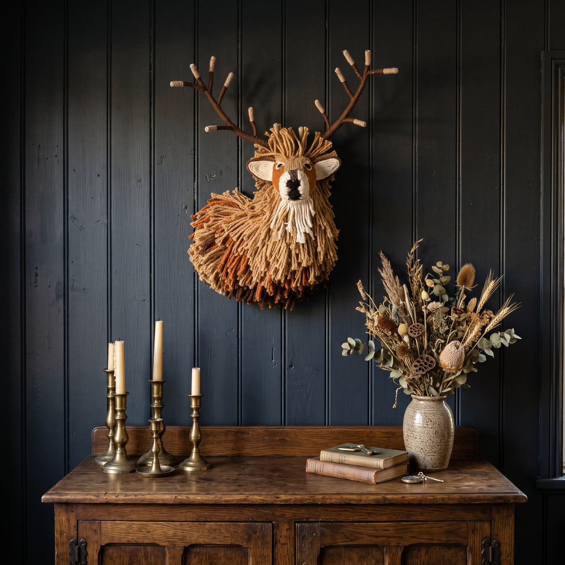 A rustic deer head wall mount with antlers and a shaggy tan fur-like mane, displayed on a dark paneled wall above a wooden console table with candles, books, and a vase of dried flowers.