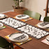 A dining table set with rectangular placemats featuring a bold black and white leaf pattern, styled with white plates and orange napkins in a warm, modern dining room setting.