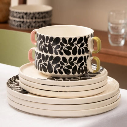 A stack of cream-colored ceramic tableware, including plates and bowls, decorated with a bold black leaf pattern. Two matching cups with pastel pink and yellow handles sit on top of the stack, set against a softly blurred indoor background.