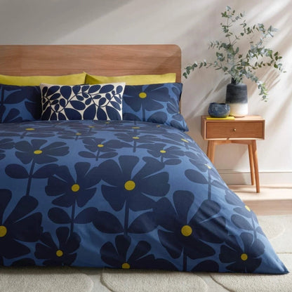 A bed dressed in a blue floral duvet cover and matching pillow shams with dark navy and bright yellow flowers, styled in a bright bedroom with a wooden headboard, side table, and plant.