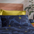 A styled bed featuring a navy blue and blue floral pattern duvet cover and matching pillowcases with yellow flower centers, layered with solid yellow pillows, set against a wooden headboard and a plant.