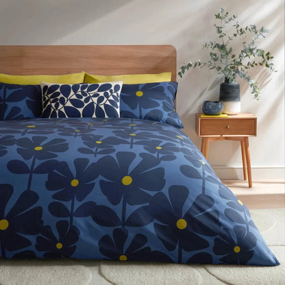 A bed dressed in a blue floral duvet cover and matching pillowcases, styled in a bright bedroom with a wooden headboard, side table, vase of greenery, and warm sunlight.