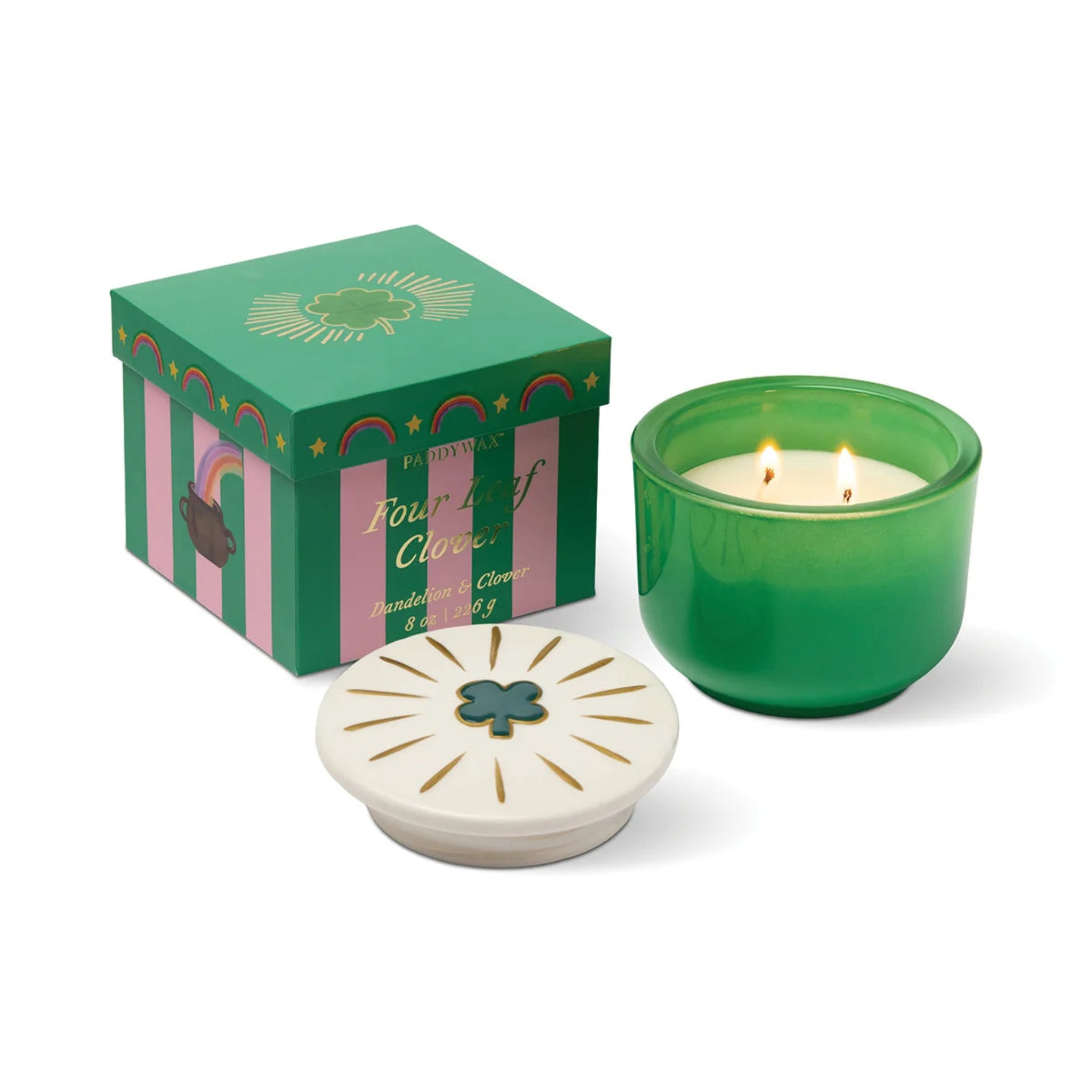 A green glass candle jar with two lit wicks sits beside its decorative green and pink gift box and a white lid featuring a green clover emblem, on a clean white background.
